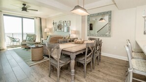 Uma sala de jantar com mesa de madeira, cadeiras e luminária pendente. Há uma varanda com cadeira e vista para o exterior.