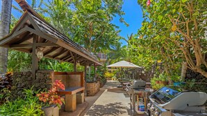 Ein Terrassenbereich im Freien mit einer Steinwand, Holzmöbeln, einer Grillstation und üppiger tropischer Bepflanzung.
