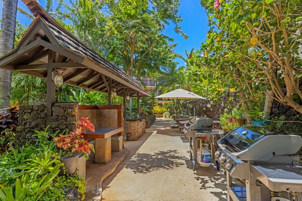 An outdoor patio area with a stone wall, wooden seating, a grill station, and lush tropical vegetation.