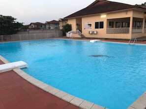 A swimming pool with a tiled deck, a covered area with a roof, and a building with a beige facade and a brown roof. - The Villa 9 at Seaview Estate (Freetown)