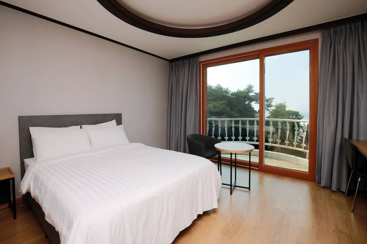 Hotelkamer met een groot bed, een klein rond tafeltje, een stoel en een balkon met uitzicht op bomen en een hekwerk.