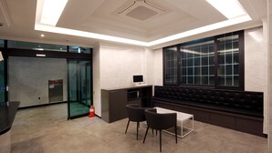 Een moderne hotelkamer met een glazen lift, een bureau met computer, een zithoek met een gestoffeerde bank en een groot raam met een rasterpatroon.