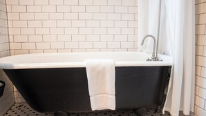 Une baignoire noire sur pieds, avec une serviette blanche, un lavabo blanc et un rideau de douche, dans une salle de bain aux murs et au sol recouverts de carreaux blancs.