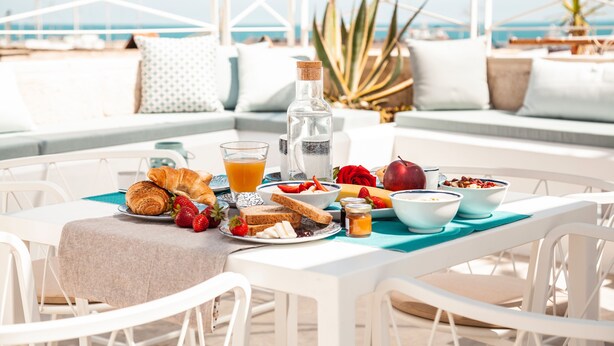 Una mesa de desayuno en una terraza con vista al mar, con croissants, fruta y una botella de jugo.