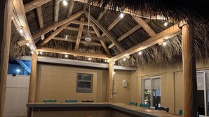 A covered outdoor bar area with wooden beams, a thatched roof, and a brick floor.