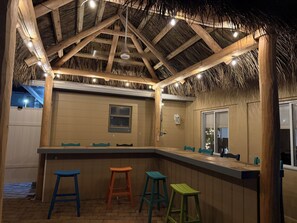 A covered outdoor bar area with wooden beams, a thatched roof, and a brick floor.