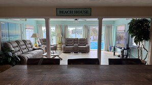 A living room with a sofa, two armchairs, a coffee table, and a TV. A sign above reads "BEACH HOUSE".