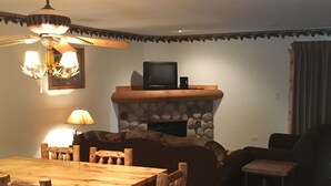 Un salon chaleureux avec une cheminée en pierre, une table à manger en bois entourée de chaises, un téléviseur posé sur la cheminée et une lampe sur la table d’appoint.