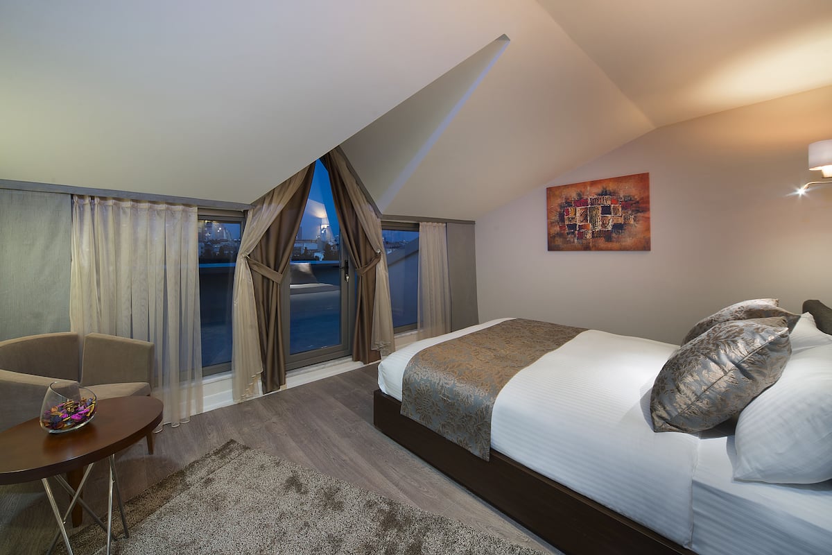 A modern hotel room with a large bed, a seating area with a small table, and a view of the city through the window.