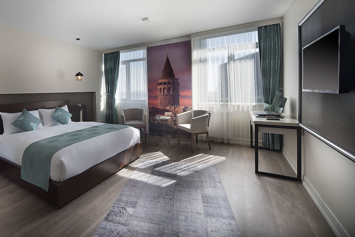 A modern hotel room with a large bed, a desk with a laptop, and a view of a cityscape through the window.