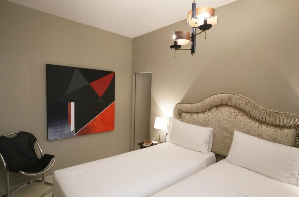A hotel room with two beds, a modern headboard, a wall-mounted abstract painting, a chair, and a bedside table with a lamp.