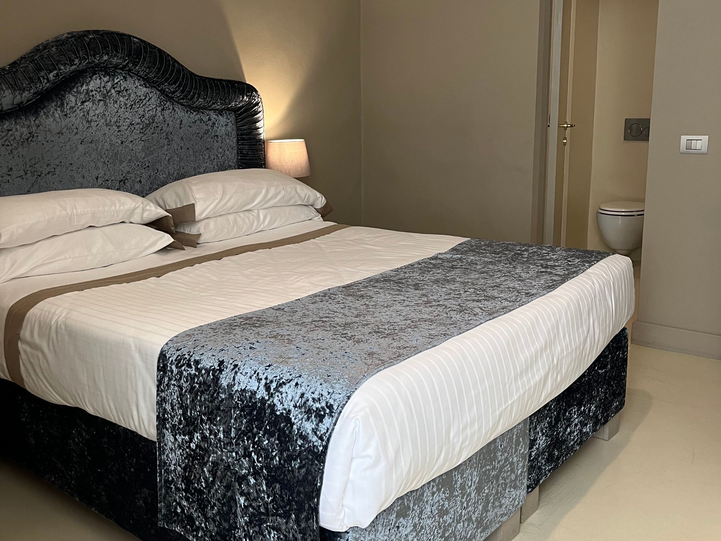 Una cama bien tendida con un cabecero oscuro, ropa de cama blanca y una manta gris estampada.