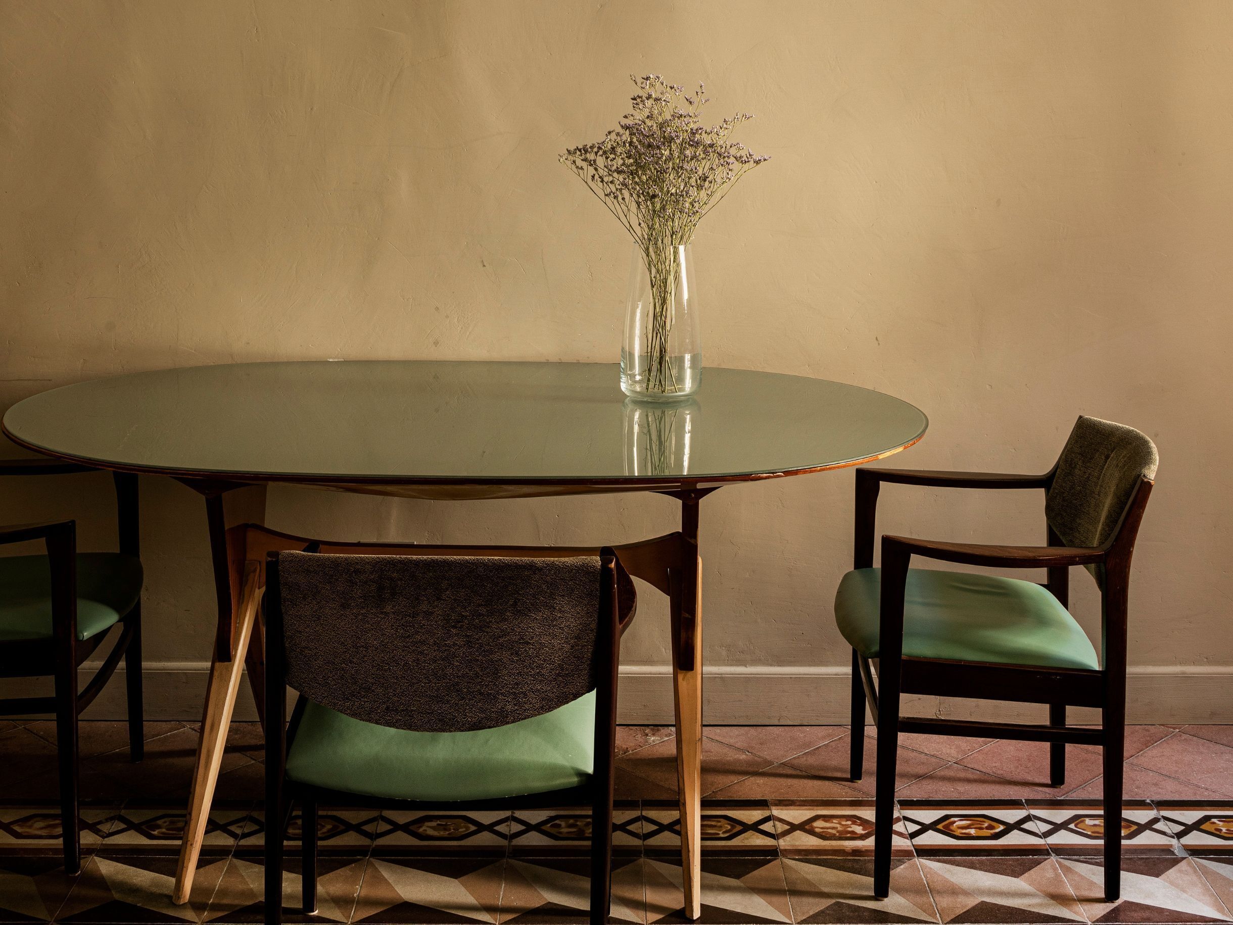 A round glass table with a vase of dried flowers, surrounded by two chairs with green upholstery.