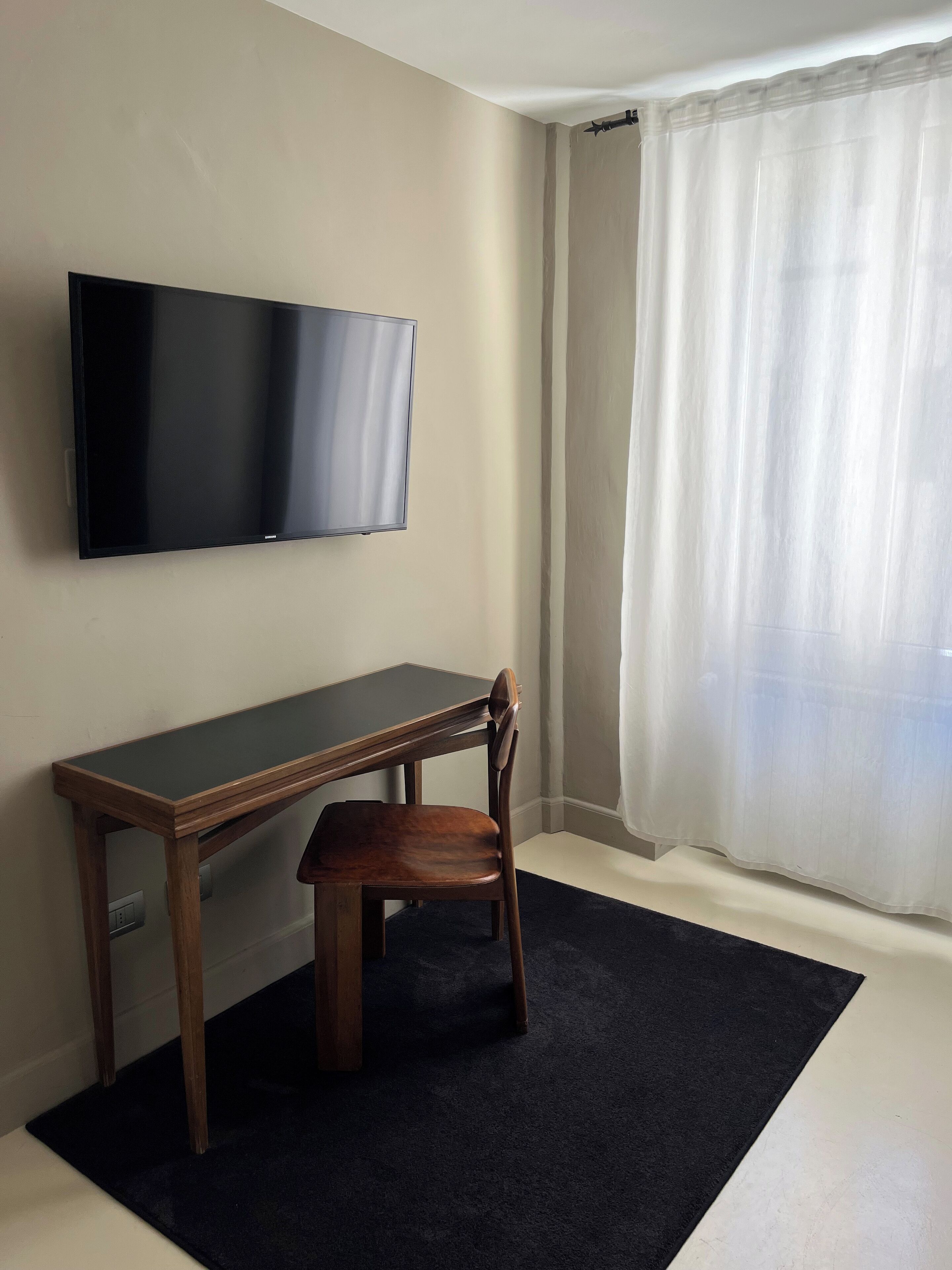 A room with a flat-screen TV mounted on the wall, a wooden desk with a chair, and a large window with sheer curtains.