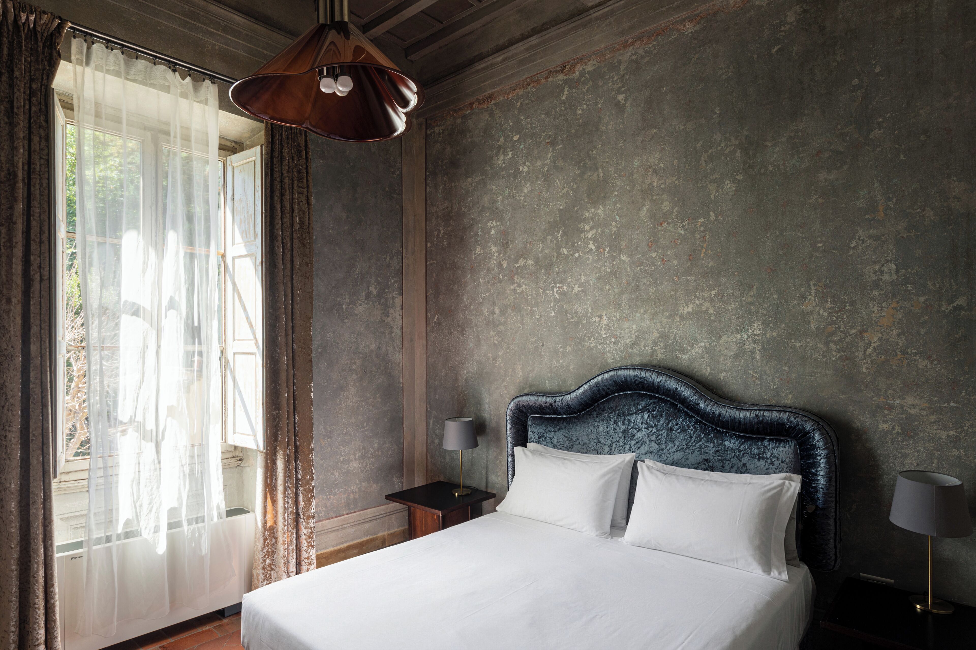 A bedroom with a large bed, a nightstand, a lamp, and a view through sheer curtains.