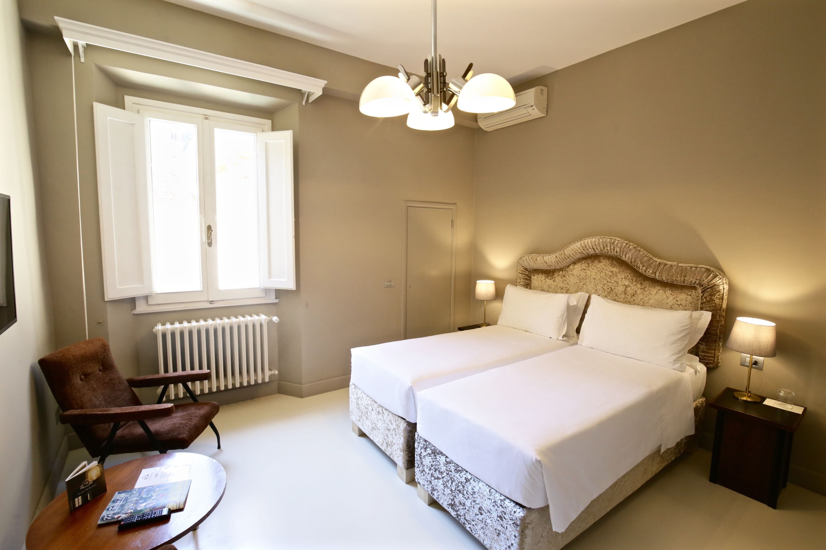 Ein Hotelzimmer mit Doppelbett, einem Sessel, einem kleinen Tisch und einem Fenster mit geöffneten Rollläden.
