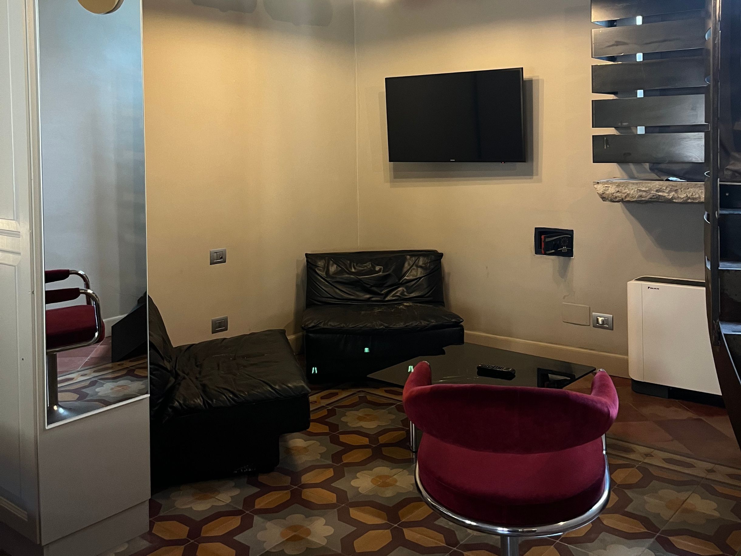 Un espace de vie compact comprenant un canapé, un fauteuil rouge, un téléviseur à écran plat fixé au mur et un petit réfrigérateur.