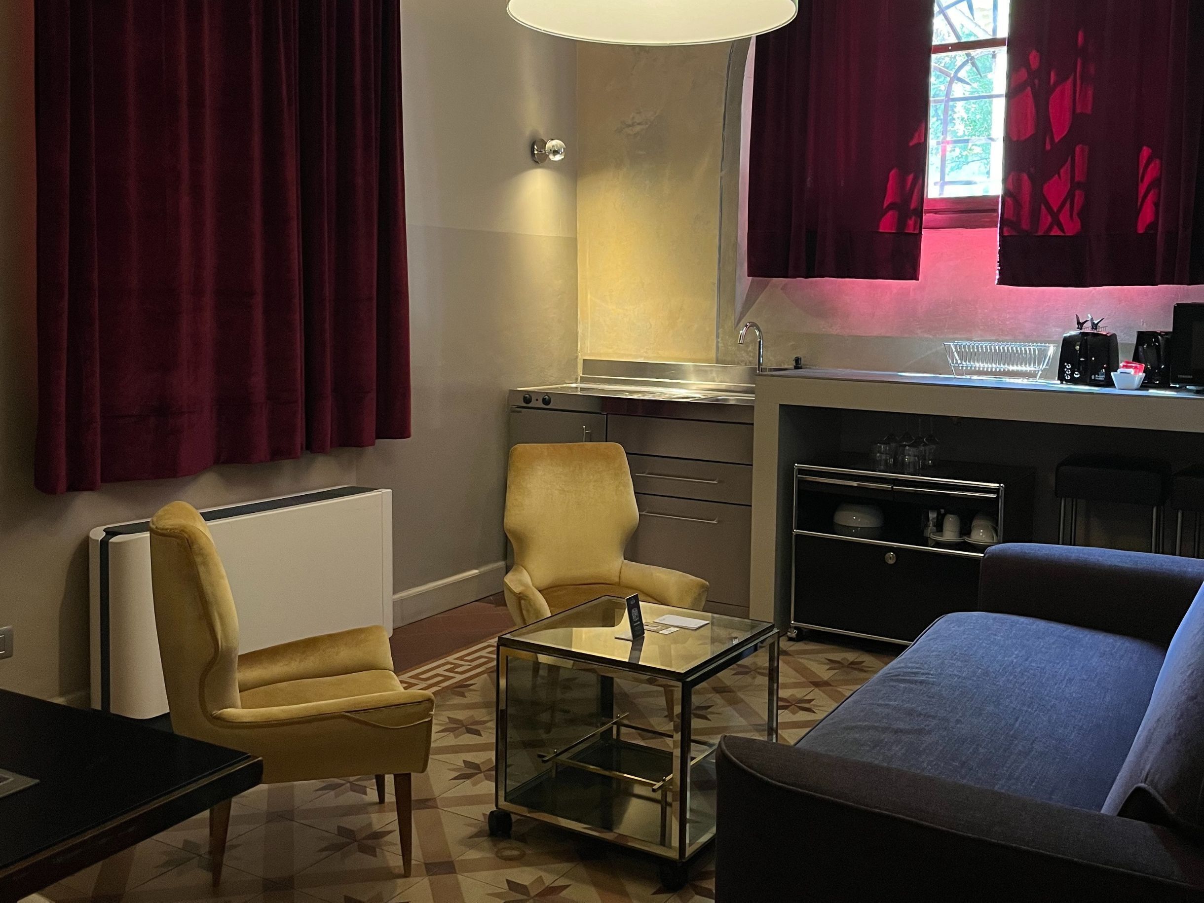 A compact living space with a sofa, armchair, and a glass coffee table. A kitchenette with a stove and sink is visible, along with a window with red curtains.