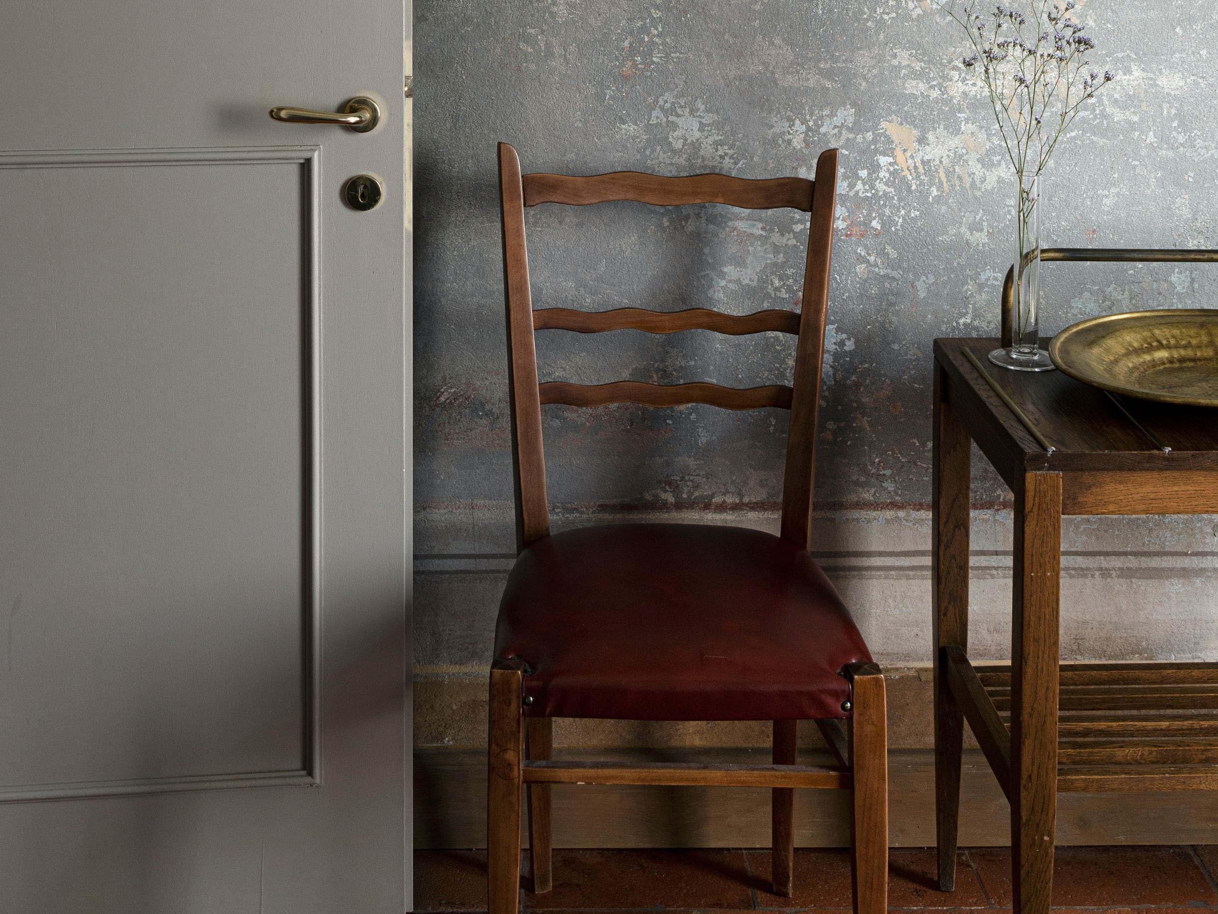 A wooden chair with a cushioned seat and backrest, positioned in front of a rustic wall with peeling paint.