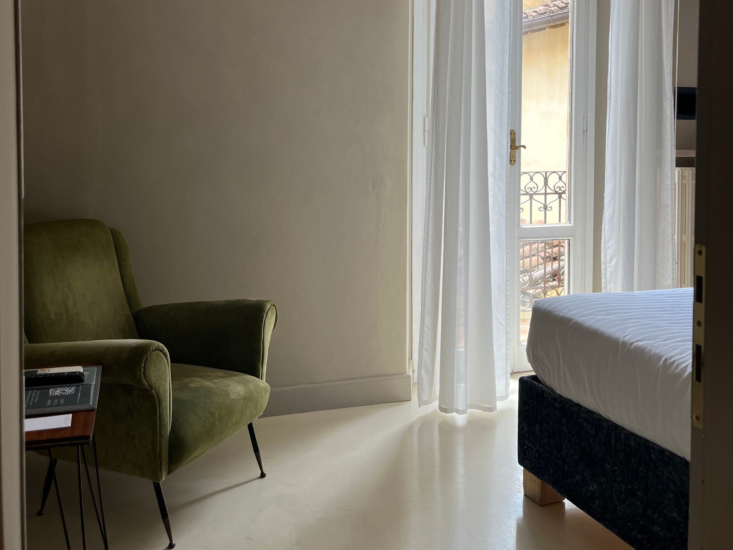 A hotel room with a green armchair, a bed with a dark headboard, and a view of a balcony through sheer curtains.
