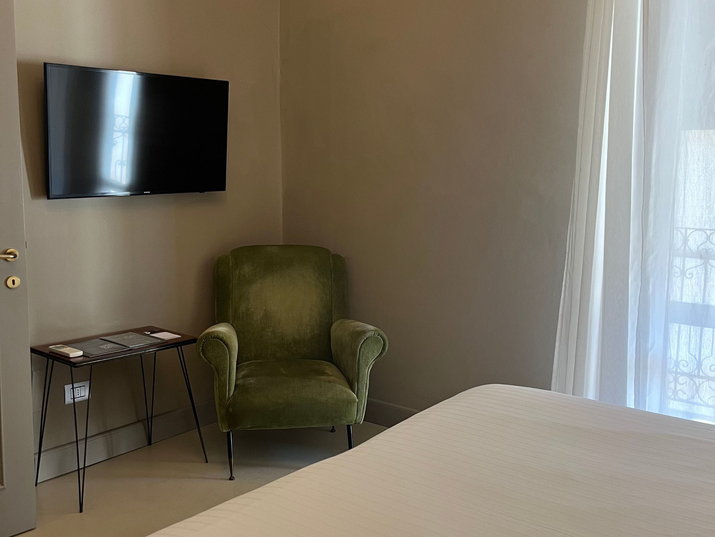 Une chambre d’hôtel avec un lit, un fauteuil vert, une petite table avec une lampe et un téléviseur à écran plat fixé au mur.