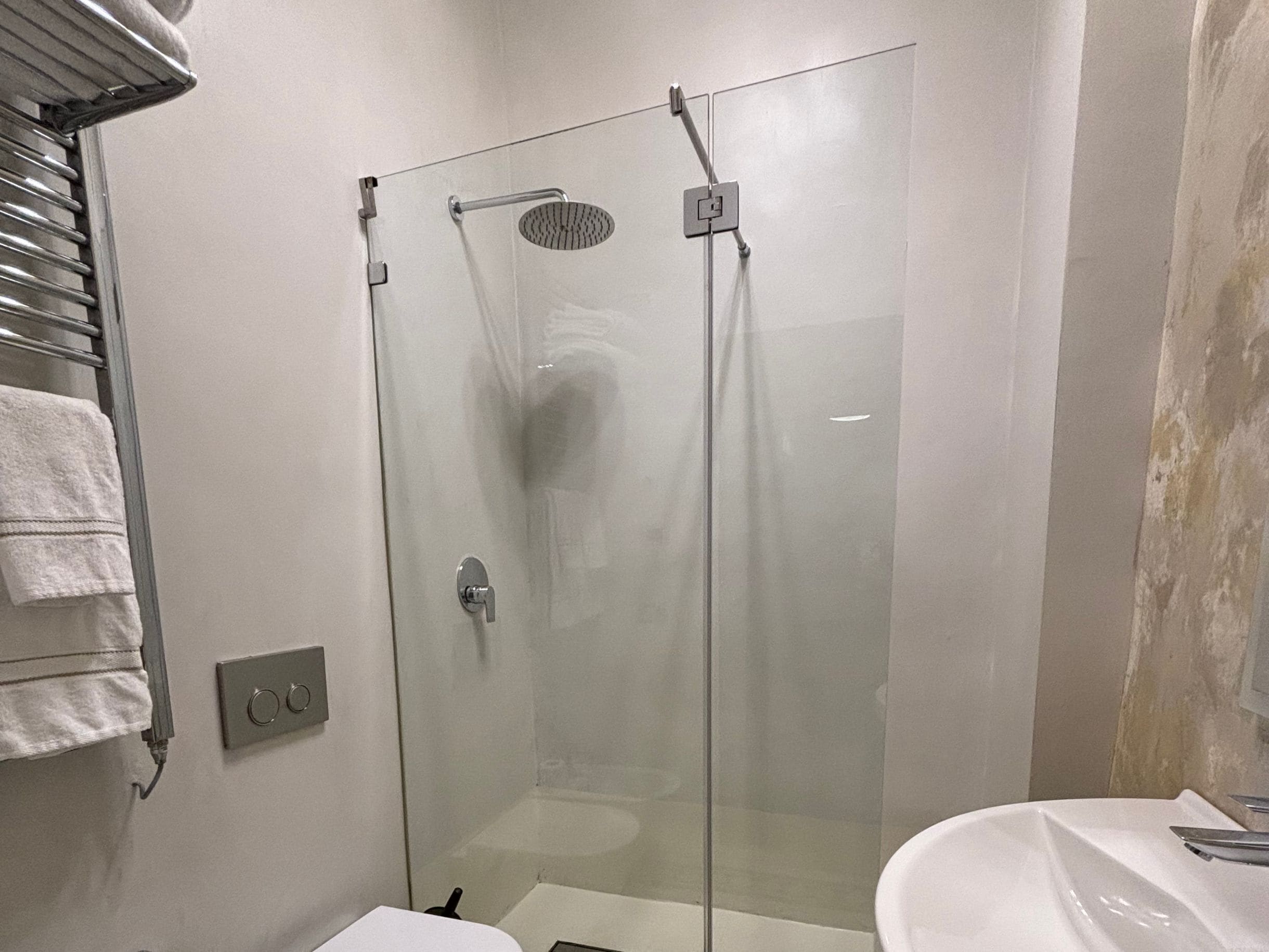 En dusch med glasväggar, ett handhållet duschmunstycke, en väggmonterad kontrollpanel och en vit tvättställ med spegel.