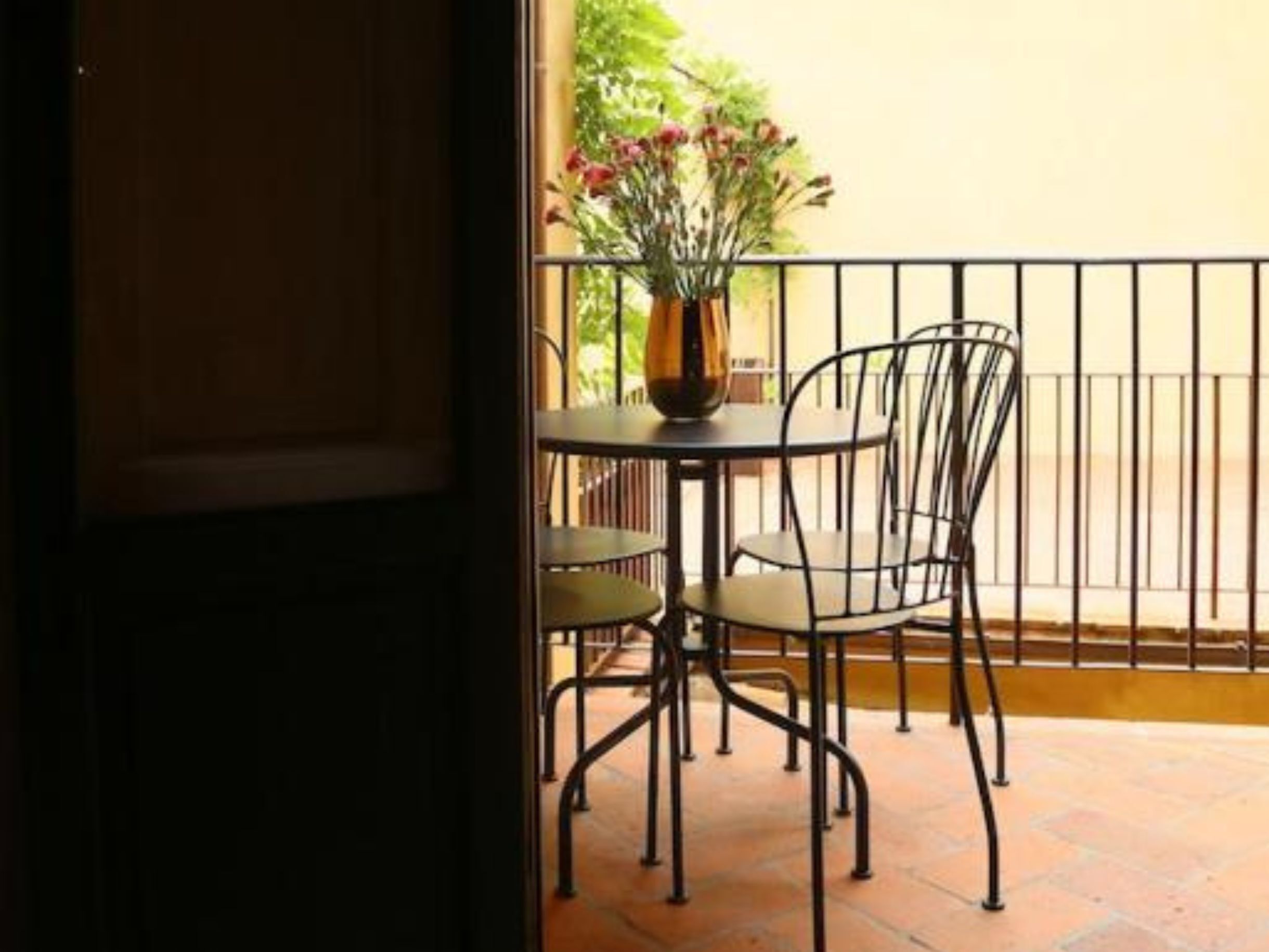 Una veranda esterna con un tavolo rotondo e due sedie, un vaso con fiori e una ringhiera in metallo.