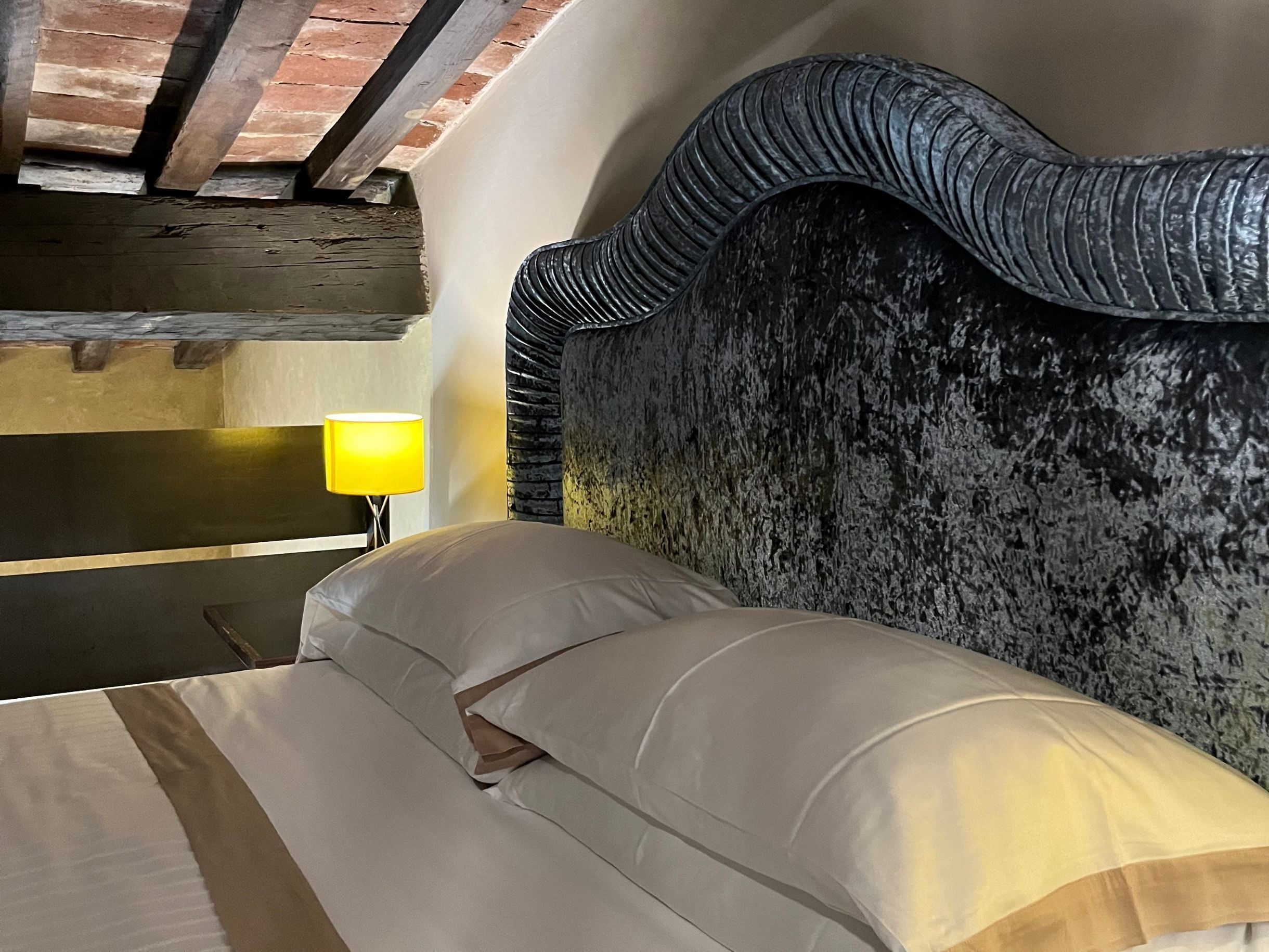 Una cama con cabecera texturizada, una lámpara de noche y una pared de ladrillo.