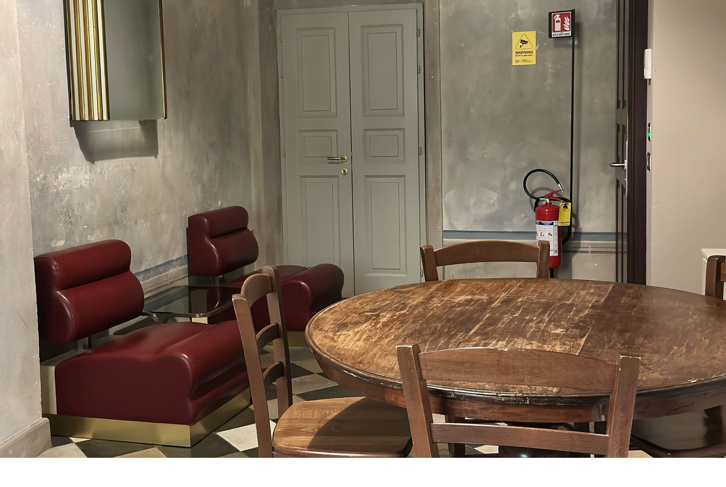 A room with a round wooden table, red upholstered chairs, a fire extinguisher, and a closed door.