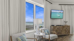 Hotelkamer met een groot raam met uitzicht op de zee en een weg. Er is een televisie aan de muur, een tafeltje met een lamp en twee stoelen met kussens.