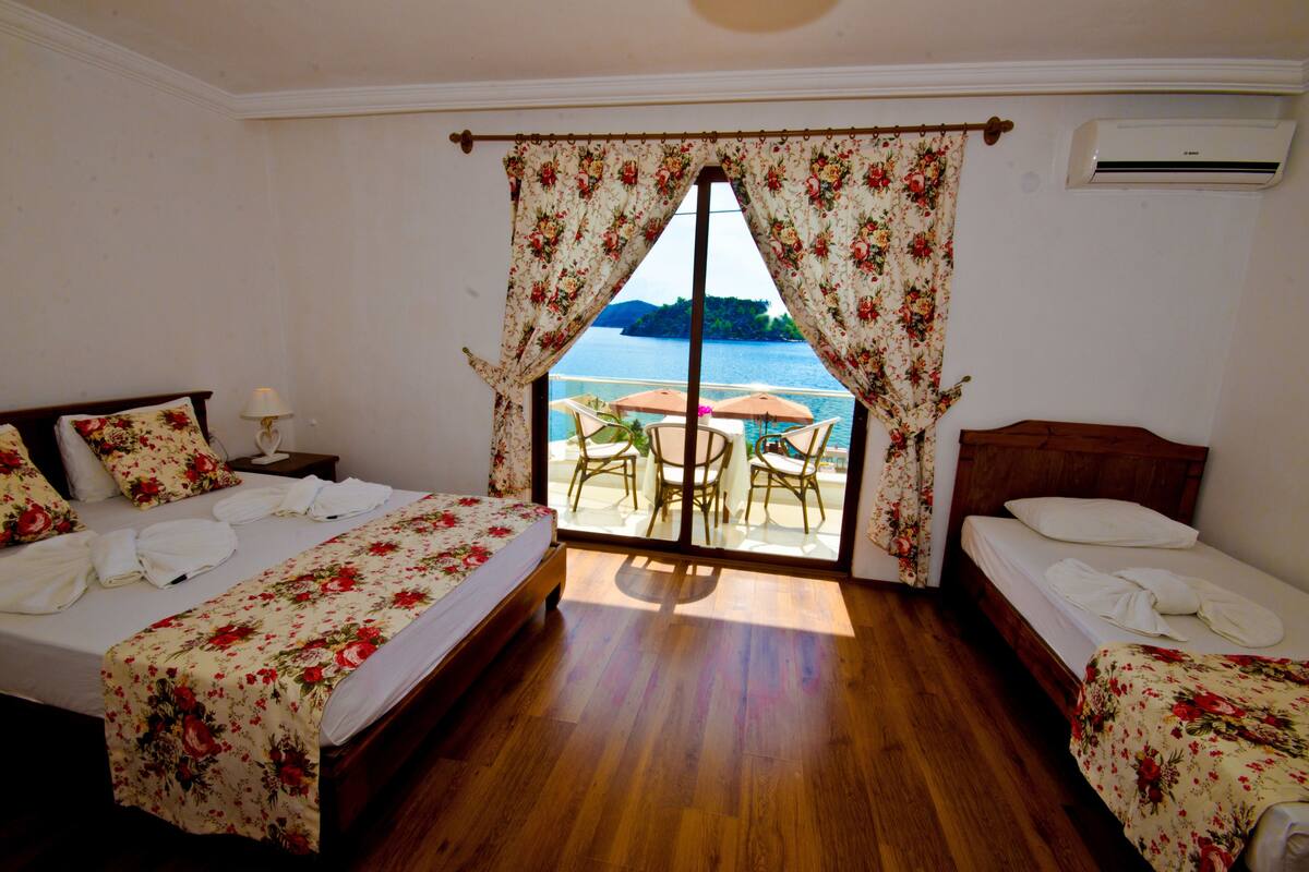 Quarto de hotel com duas camas, uma varanda com mesa e cadeiras, e vista para o mar.