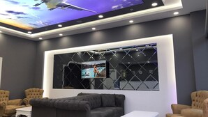 Eine moderne Lounge mit großem Fernseher, der ein Flugzeug zeigt, einer Couch, Sesseln und einem Couchtisch.