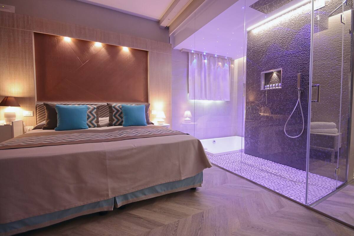 A modern hotel room with a large bed, a glass-enclosed shower area, and a view of the cityscape.