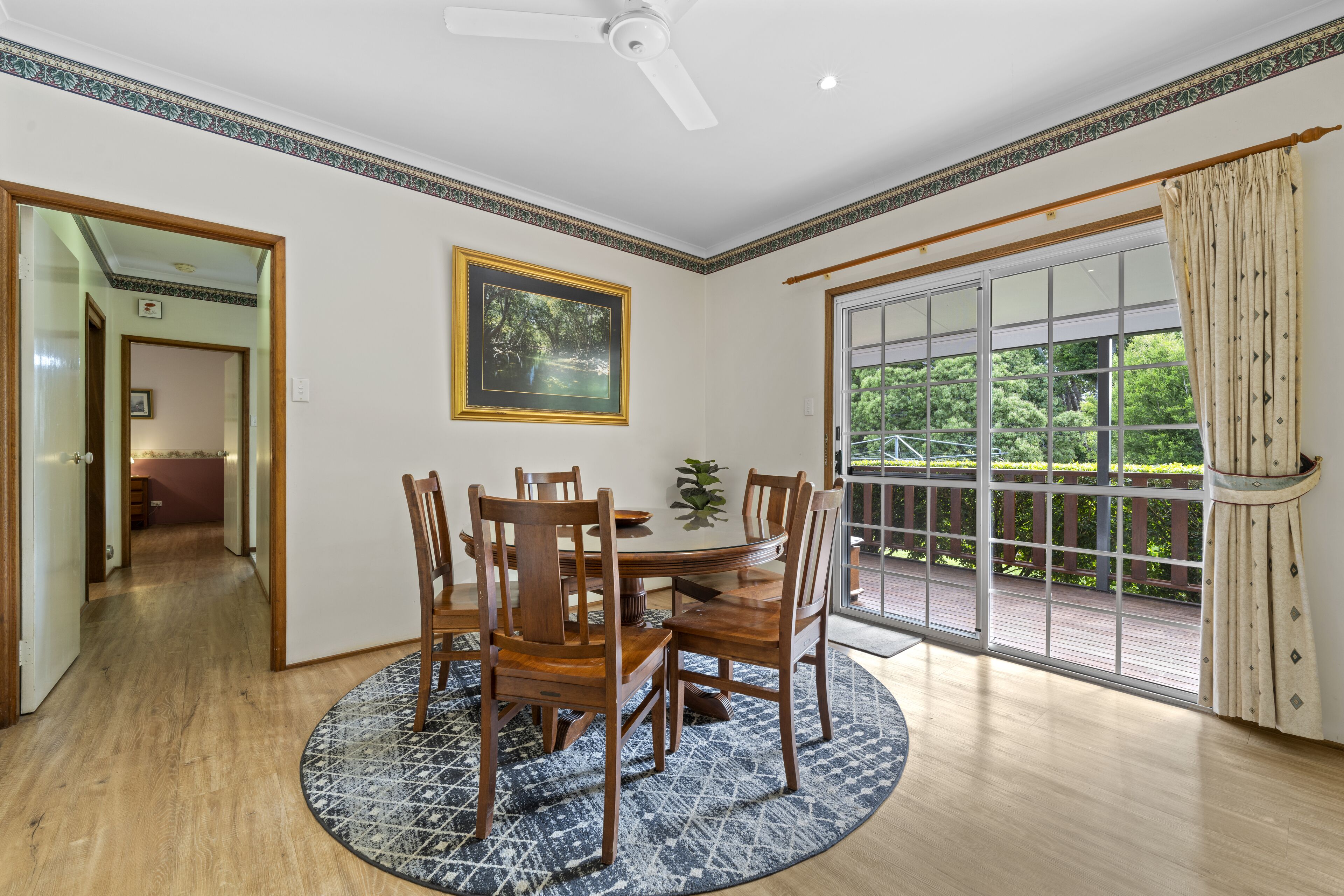 A dining area with a round table, wooden chairs, a ceiling fan, and a large window with a view of greenery.