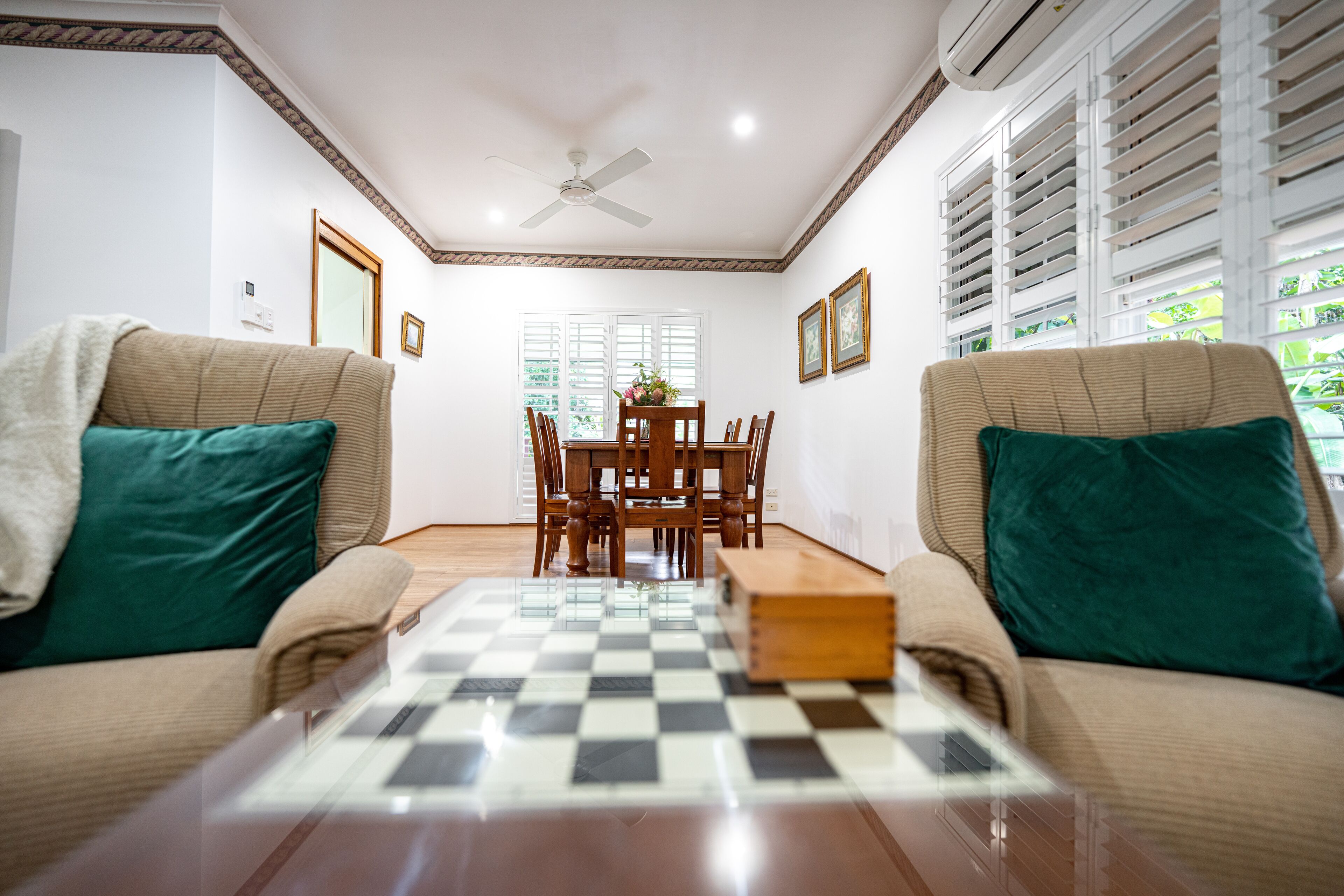 A room with a checkered floor, two beige sofas with green cushions, a wooden coffee table, a dining table with chairs, and a ceiling fan.
