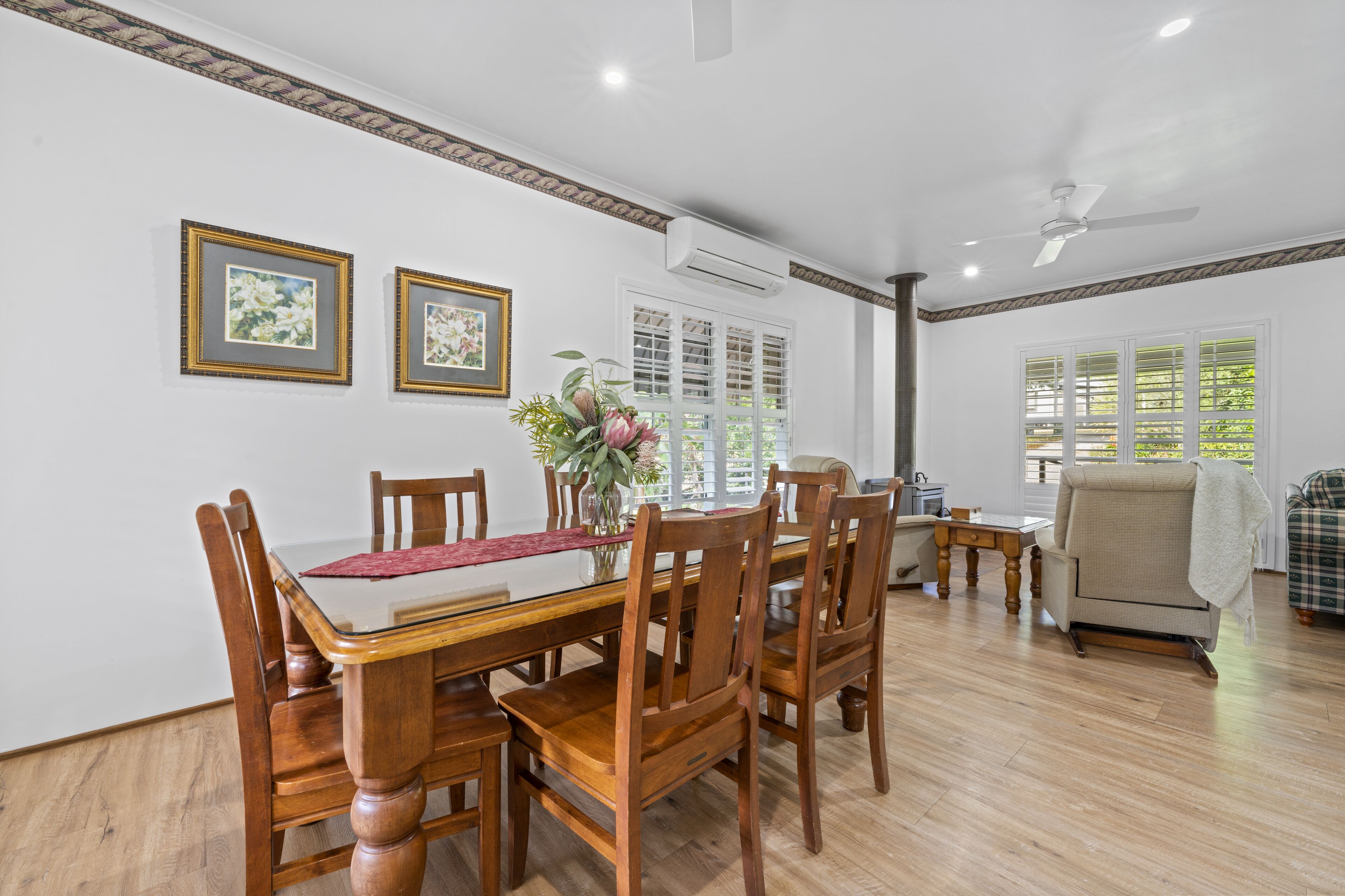 A dining area with a wooden table and chairs, framed pictures on the wall, and a window with blinds.
