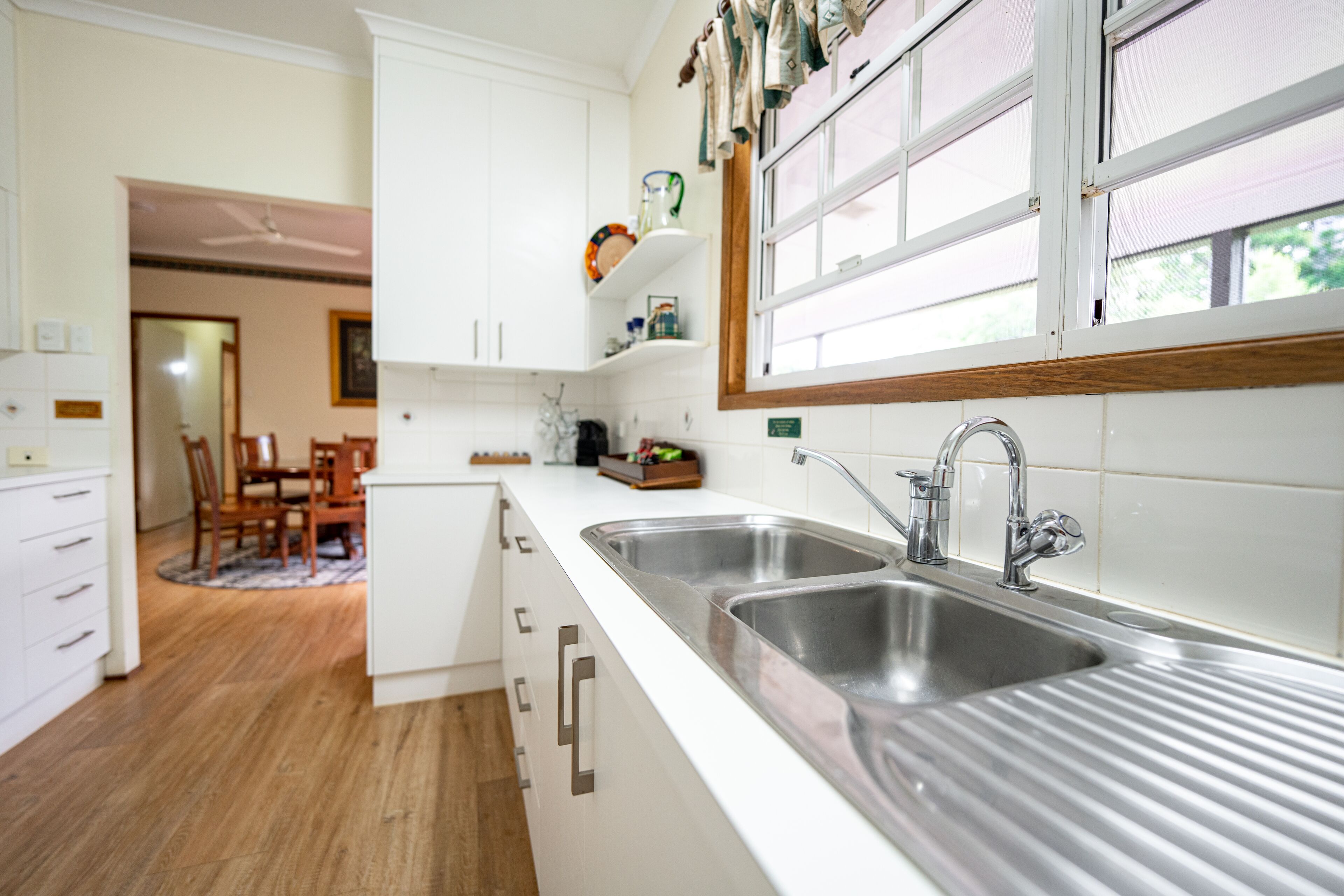 A modern kitchen with white cabinetry, stainless steel sinks, and a wooden window frame.