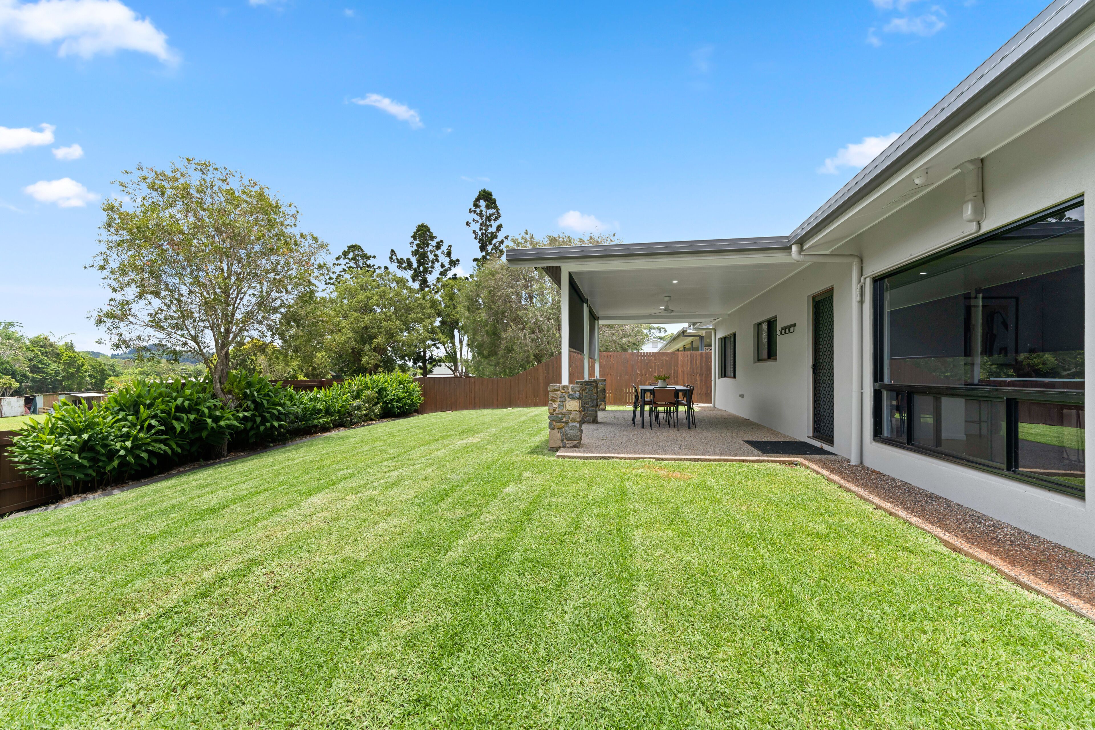 A modern house with a well-maintained lawn, a patio with a table and chairs, and a large window.