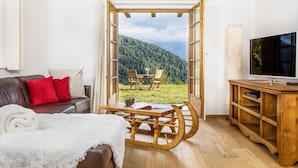 Uma sala de estar com uma mesa de madeira, um sofá com almofadas vermelhas, uma TV sobre um suporte de madeira e vista para uma encosta arborizada através de uma porta aberta.