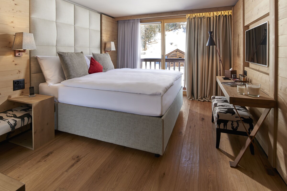 Ein modernes Hotelzimmer mit einem großen Bett, Holzboden, einem Schreibtisch mit Stuhl und einem Vorhangfenster.