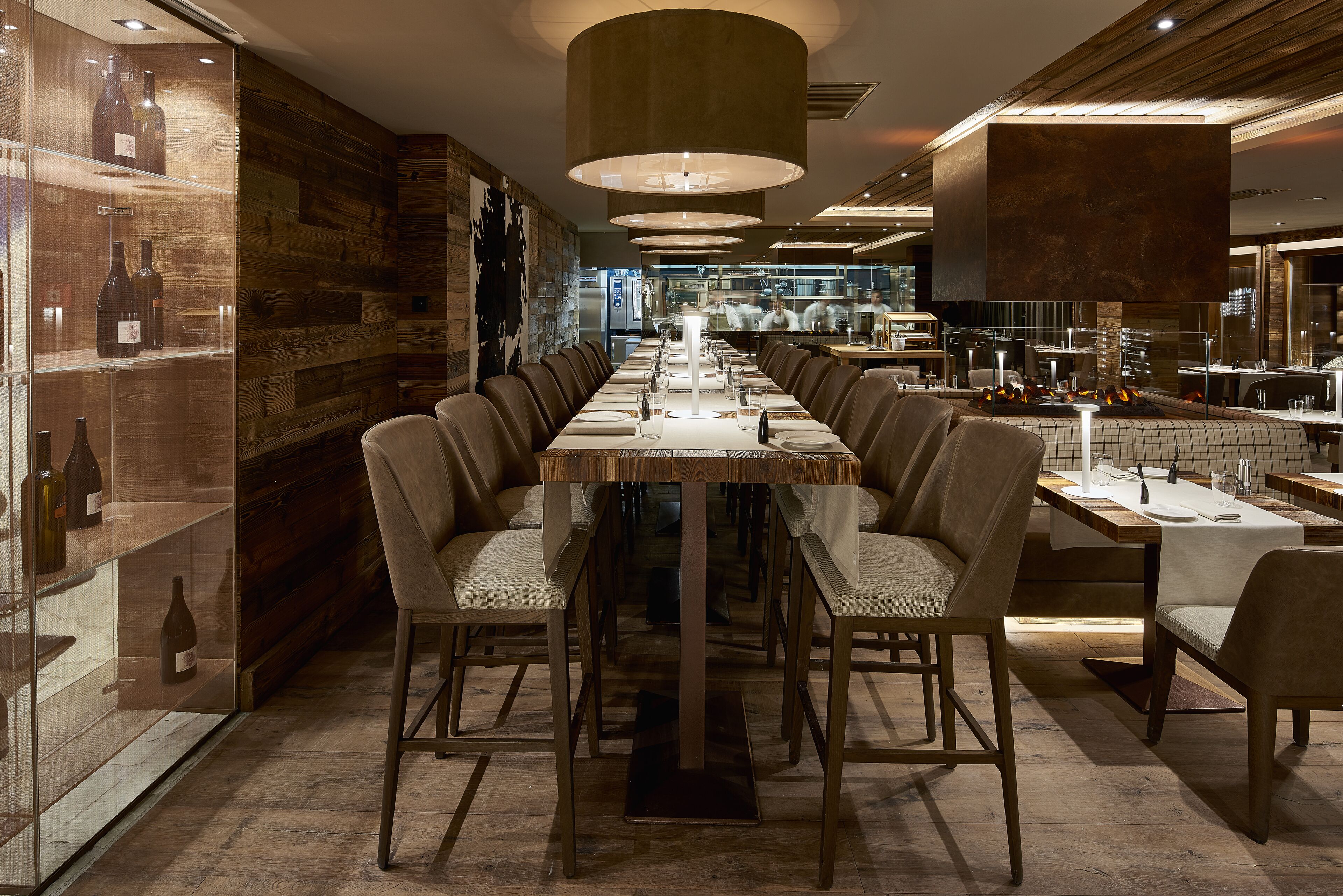 A long dining table with multiple chairs, a wine display, and a modern kitchen in the background.