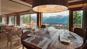 Uma área de refeições com mesa de madeira posta para dois, grandes janelas com vista para a montanha e um lustre.