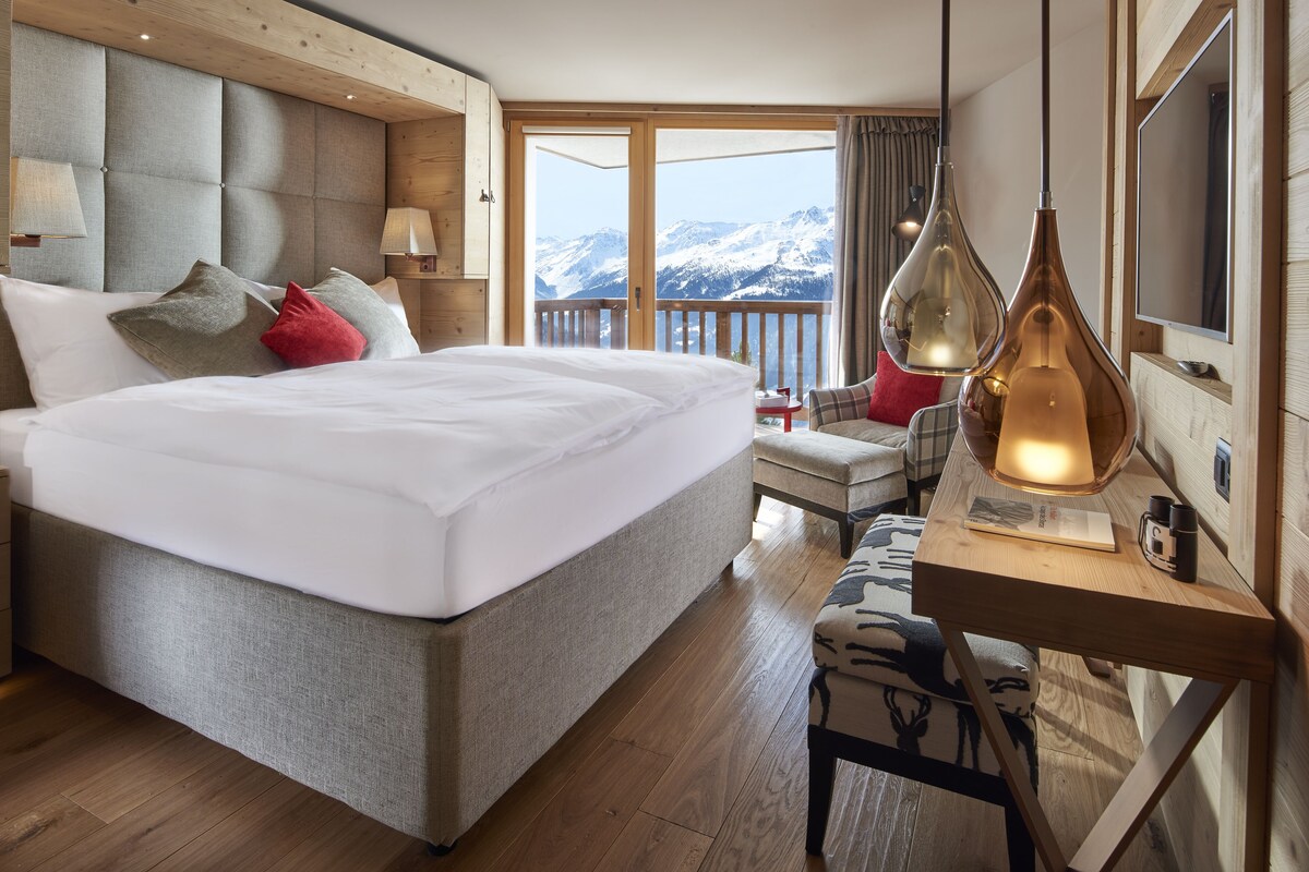 Ein Hotelzimmer mit einem großen Bett, einem Schreibtisch mit Lampe und Blick auf schneebedeckte Berge.