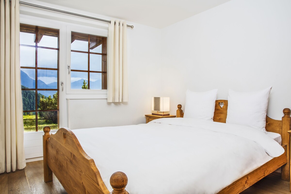 Ein Schlafzimmer mit einem hölzernen Bett, einem Nachttisch, einer Lampe und einem Fenster mit Vorhängen.