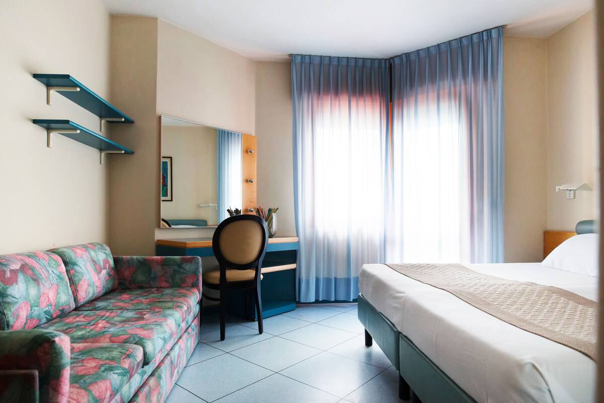 A hotel room with a bed, a desk, a chair, a sofa, and a large window with curtains.