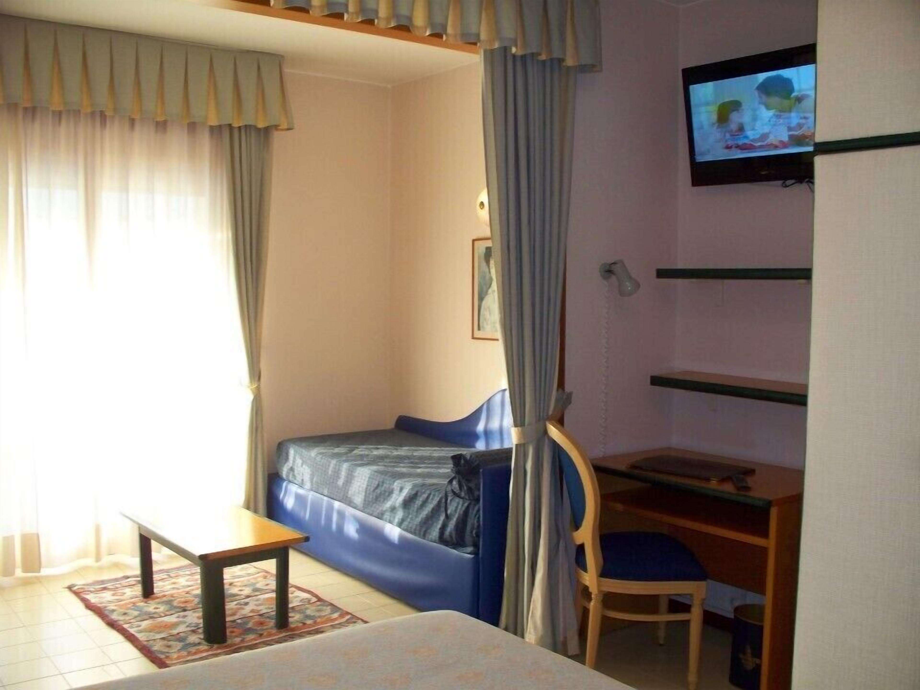 Ein Zimmer mit einem Bett, einem Schreibtisch mit Stuhl, einem Fernseher und einem Fenster mit Vorhängen.
