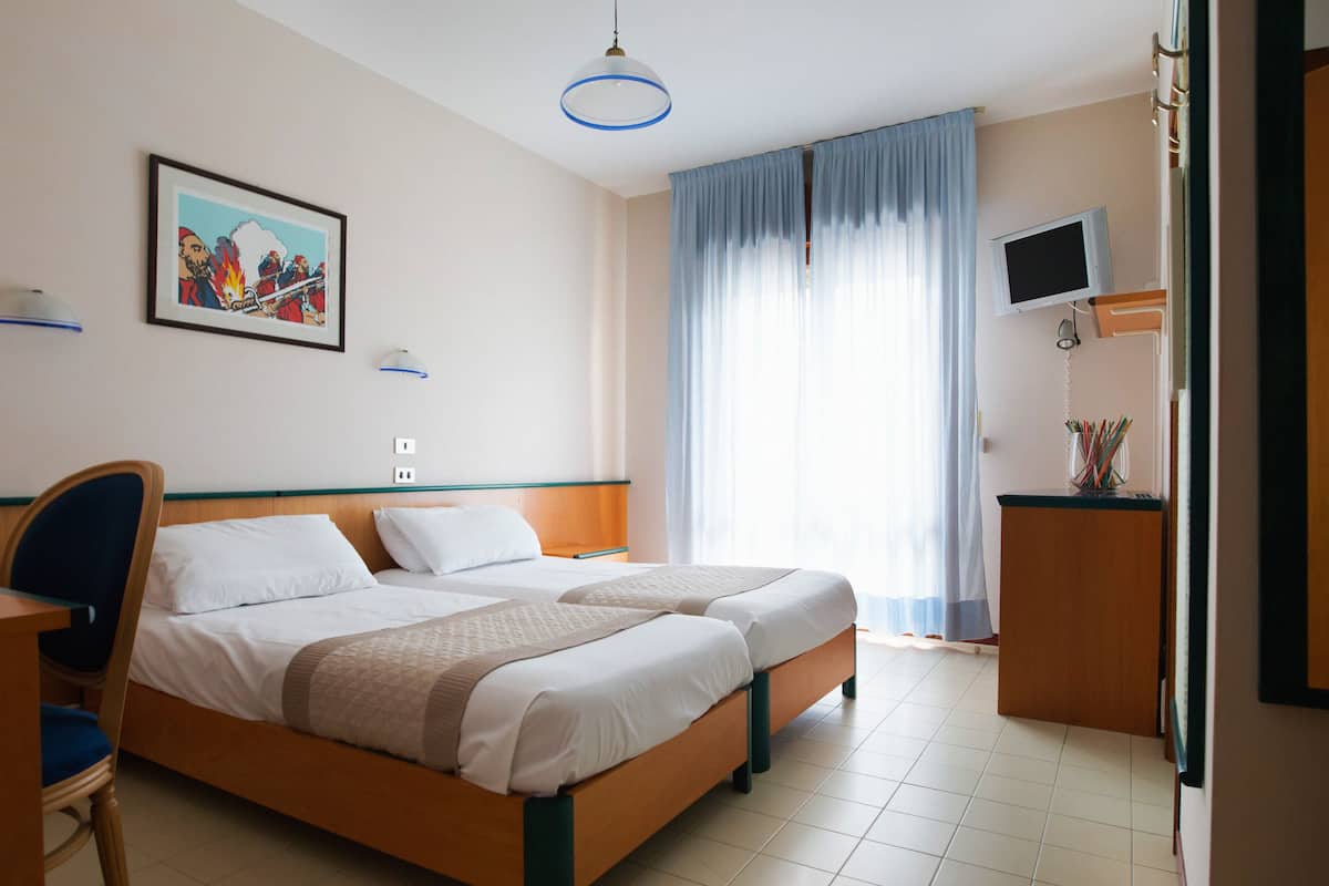 A hotel room with two beds, a desk, a chair, a TV, and a painting on the wall.