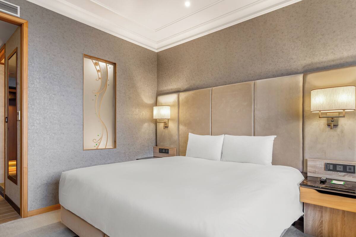 A hotel room with a large bed, a nightstand with a lamp, a wall-mounted artwork, and a wooden door.