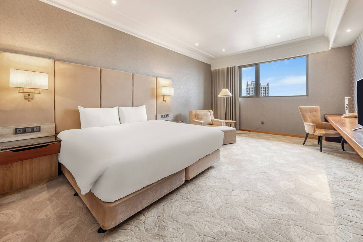 A hotel room with a large bed, bedside tables, a desk, a chair, and a window with a city view.