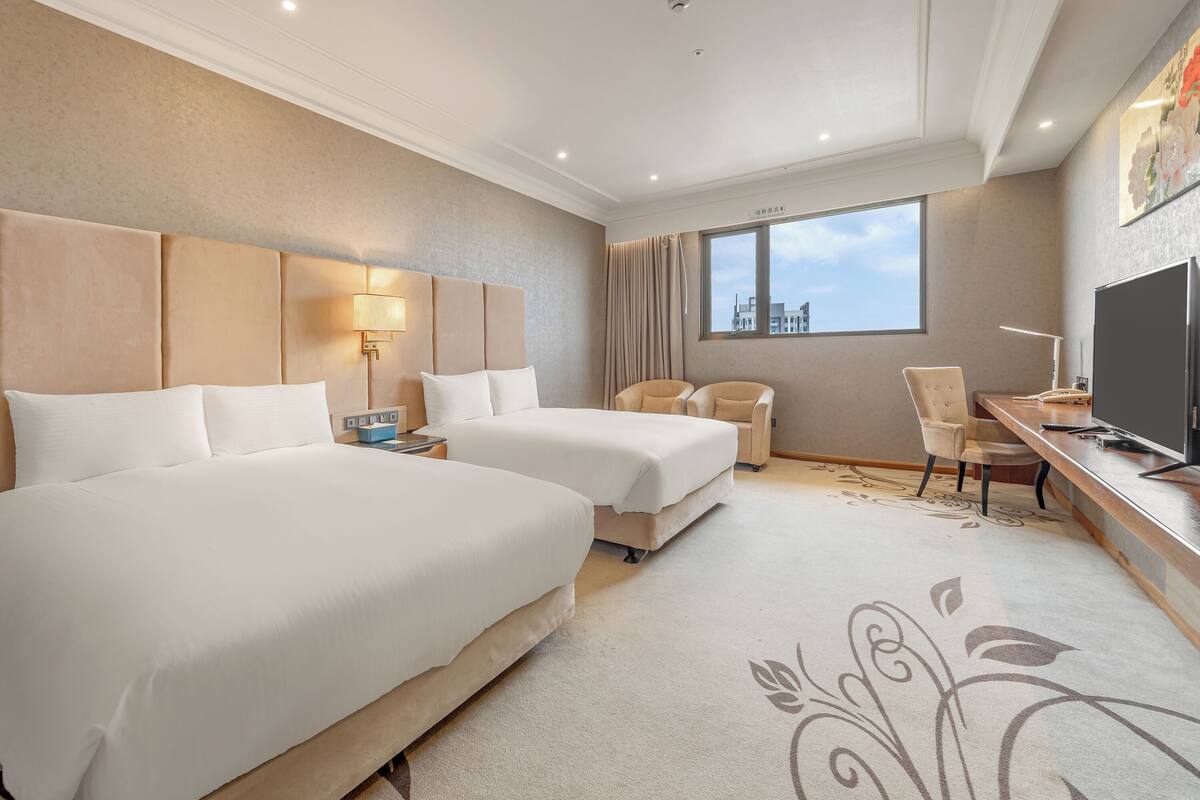 A hotel room with two beds, a desk with a TV, a chair, and a window with a city view.